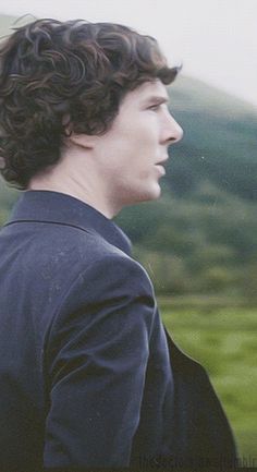 a young man with curly hair standing in front of a green field and mountain range