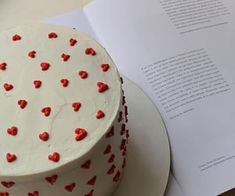 a white cake with red hearts on it next to an open book and some papers