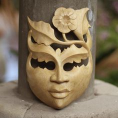 a wooden mask with flowers on it sitting on a cement pillar in front of some bushes