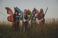 four women dressed in costumes holding swords and butterfly wings, standing in tall grass with sky background