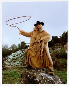 a woman in a fur coat and hat holding a lasso on top of a rock