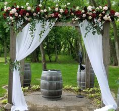 an outdoor ceremony setup with white drapes and red flowers on the arch over barrels