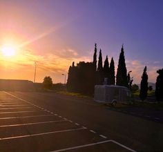 the sun is setting over an empty parking lot with trees on either side and a van in the middle