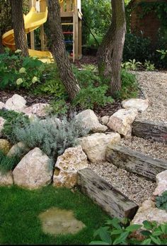 an outdoor play area with rocks, grass and trees in the foreground is a yellow slide