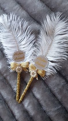 two white feathers with gold accents are laying on a fur surface, one has a name tag and the other has a bow