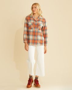 a woman standing in front of a wall wearing white pants and an orange plaid shirt