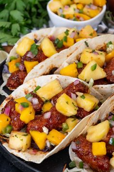 three tacos filled with meat and pineapple salsa on a black plate next to cilantro