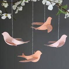 three pink paper birds hanging from strings with white flowers in the background and gray wall