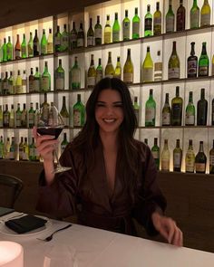 a woman sitting at a table with a glass of wine in front of bottles on the wall