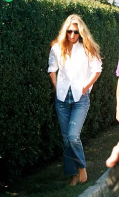 a woman in white shirt and jeans standing next to a hedge