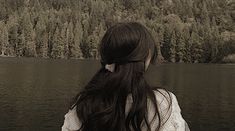 a woman with long black hair standing in front of a body of water next to trees