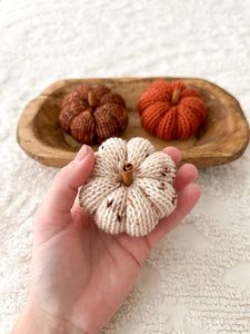 a hand is holding three knitted pumpkins in a wooden bowl on a white tablecloth