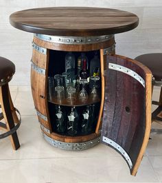 a wooden barrel table with wine glasses and liquor bottles in the bottom half, next to two stools