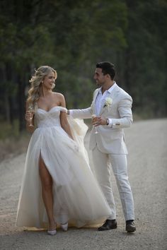 a man in a white suit and woman in a wedding dress walking down a dirt road