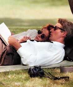 a man and woman laying on the grass reading a book under a tree in the park