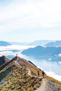 two people walking up the side of a mountain on top of a grass covered hill