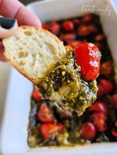 a hand holding a piece of bread with pesto and tomatoes on it in a white bowl