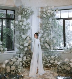 a woman standing in front of a white backdrop with lots of flowers and greenery