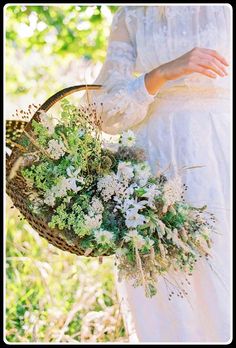 a woman holding a basket filled with flowers and greenery in her hands while standing outside