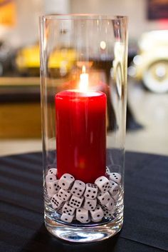 a red candle is sitting in a glass vase with dice around it on a table