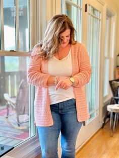 a woman standing in front of a window looking at her cell phone while wearing jeans and a pink cardigan