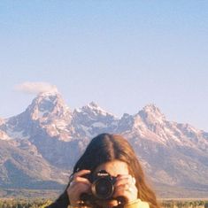 a woman holding a camera up to her face in front of the mountains with snow on them