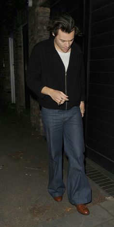 a man in black jacket and jeans walking down street at night with cell phone up to his ear