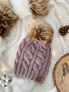 two knitted hats sitting on top of a white blanket next to scissors and pine cones