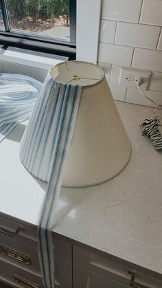 a lamp shade sitting on top of a counter next to a window with a blue and white striped ribbon