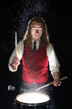 a man in a red vest and tie is playing drums with water splashing on him