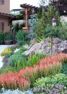 an outdoor garden with lots of flowers and plants around it, along with a house in the background