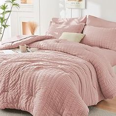 a bed with pink comforter and pillows in a room next to a plant on the floor