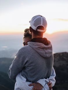 a man and woman embracing each other while standing on top of a mountain at sunset