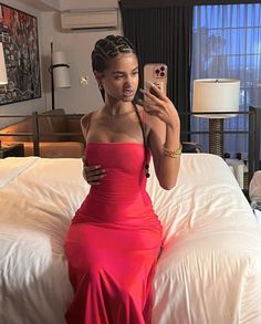 a woman in a red dress sitting on a bed taking a selfie with her cell phone