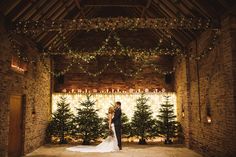 a bride and groom standing in front of christmas trees at their wedding reception with lights strung from the ceiling