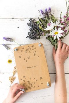 a person holding flowers next to a notebook on a white wooden table with writing in it