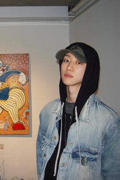 a young man wearing a denim jacket and hoodie standing in front of a painting