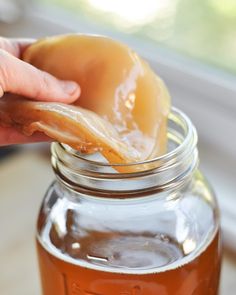 a person holding a donut over a jar filled with honey and syrup on top of a wooden table