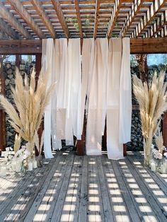 an outdoor area with white curtains and tall grass in vases on the wooden floor