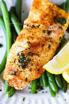 a white plate topped with fish and green beans covered in seasoning next to lemon wedges