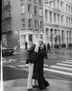 a man and woman standing on the sidewalk in front of a tall building, with their arms around each other