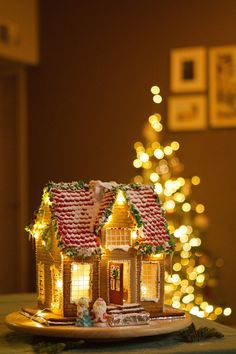 a gingerbread house on a plate with christmas lights in the background and a lit tree behind it