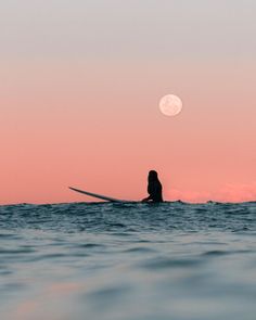 a person sitting on a surfboard in the ocean at sunset with the moon behind them