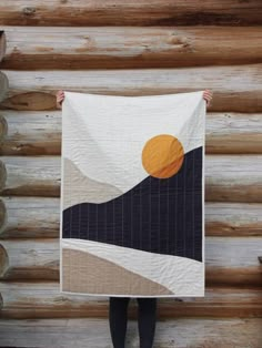 a person holding up a quilt in front of a log cabin