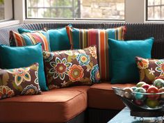 a couch with colorful pillows on it next to a bowl of fruit and a window