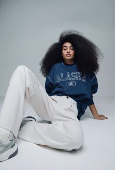 a woman with an afro sitting on the ground wearing white pants and a blue alaska sweatshirt