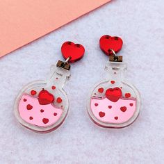 These love potions are perfect for Valentine's Day. They feature red hearts bottled in a pink elixir. There are red engraved details surrounding pink and red shiny acrylic, inside of a clear bottle with wooden corks on top. They are shiny and reflective to add a little sparkle to your life. Dimensions: 1.5 in (38 mm) L x 1.0 in (26 mm) W The drop length is about 2.0 in (51 mm). Materials: Mirror Acrylic Hypoallergenic, Nickel Free, Stainless Steel Shipping: All of our earrings are made-to-order to cut down on waste. Your earrings will be produced and shipped within a week after you order. Care: Do not soak earrings in water. Materials are water resistant, but it is not recommended that you swim, shower, or bath with your earrings on. Avoid getting lotions, oils, and perfumes on your earrin Love Potions, Valentine Earrings, Valentine Jewelry, Clear Bottle, Clay Dangle Earrings, Valentines Earrings, Love Potion, Funky Earrings, Mirror Acrylic