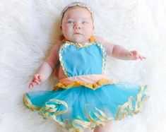 a baby wearing a blue and gold dress laying on a white furnishing with her hands out