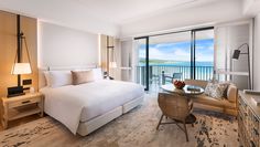 a hotel room with a bed, desk and chair in front of a balcony overlooking the ocean