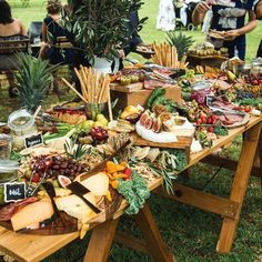 a table full of food with people standing around it in the grass near trees and bushes